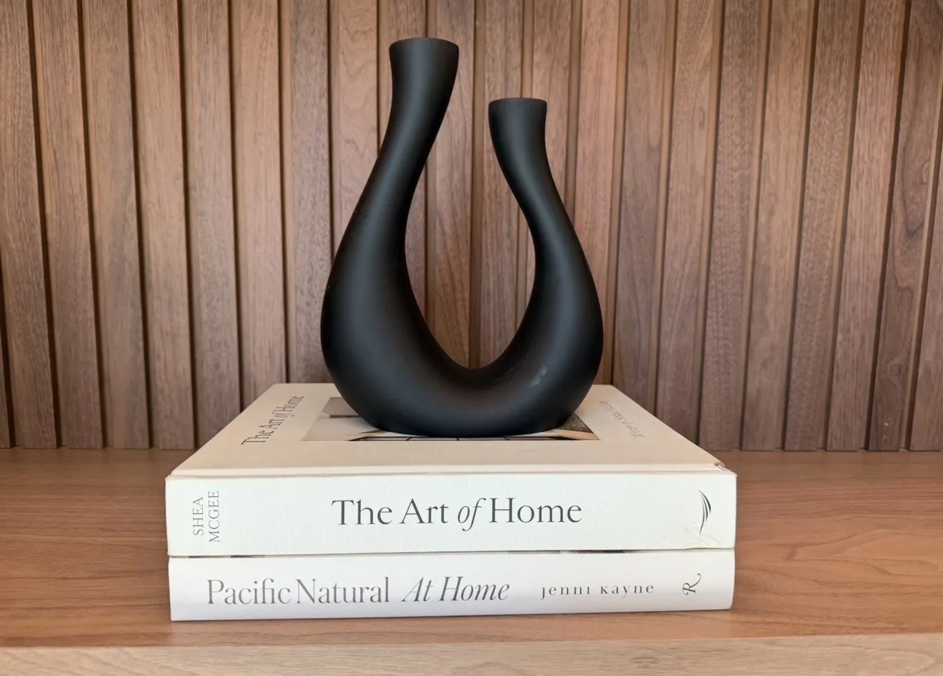 Black sculptural vase on books against a wood paneled wall