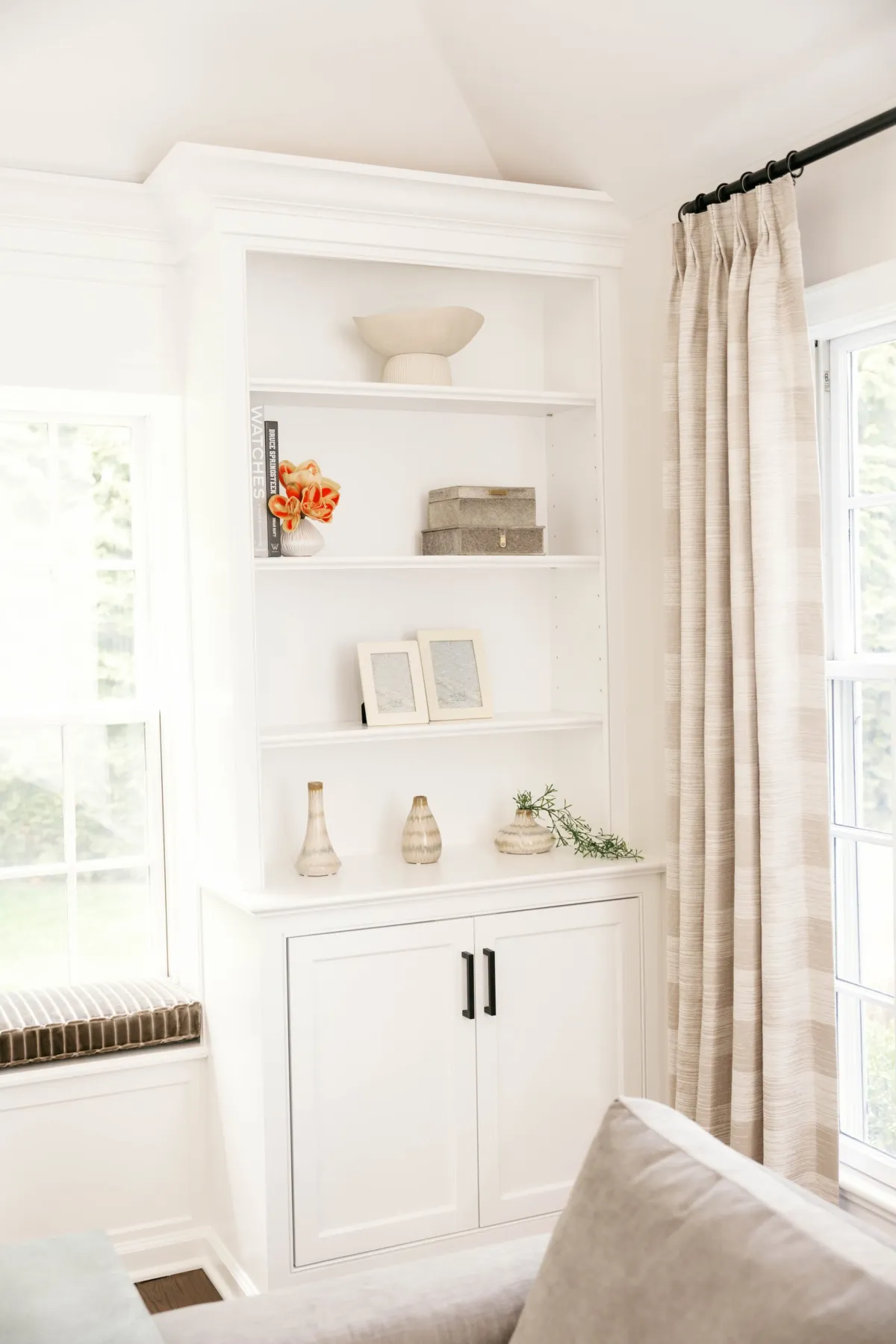 White built-in shelves styled with orange flowers, ceramics, and boxes beside tall drapery