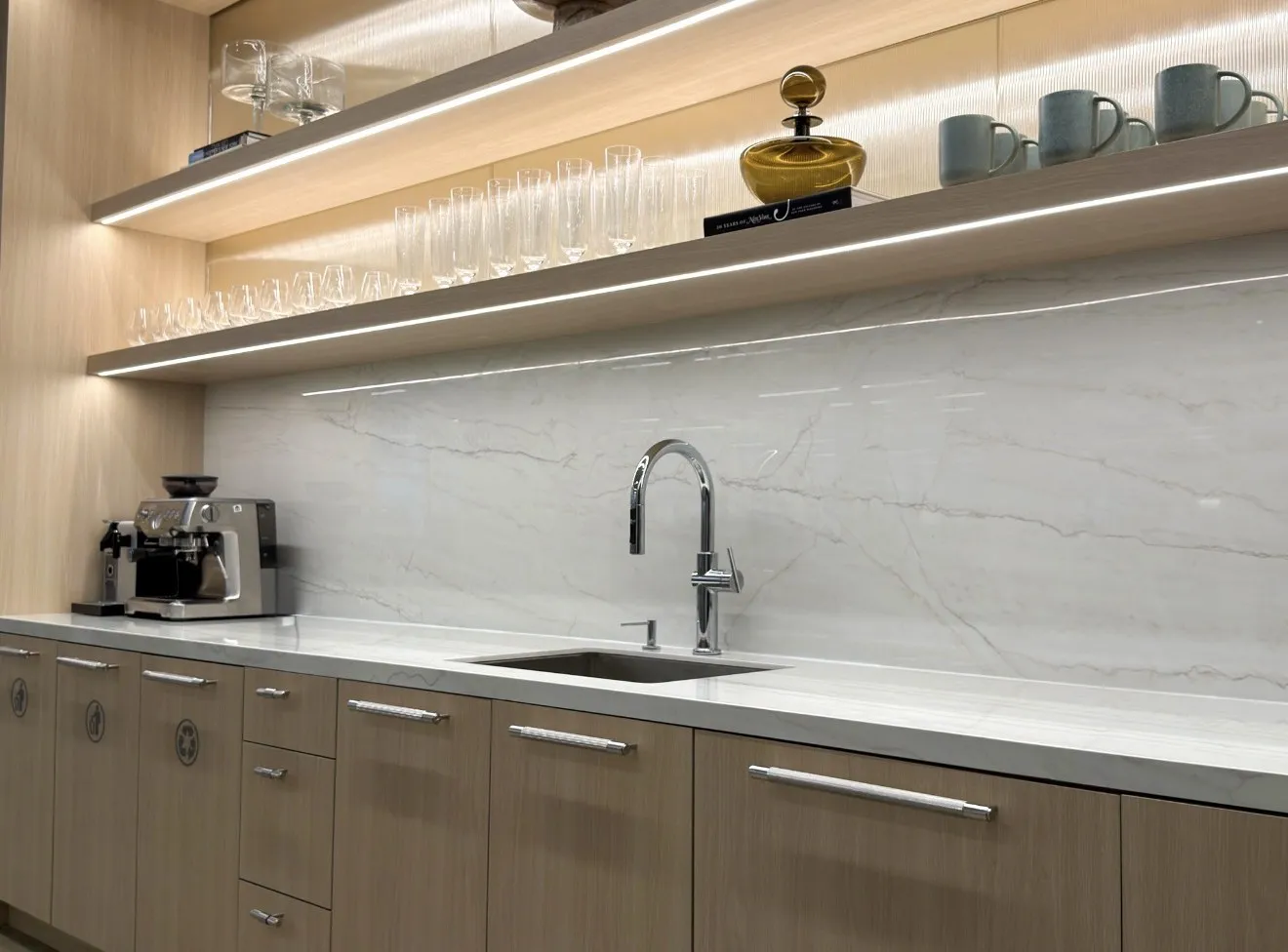 The corporate café bar seen from the side, with a marble backsplash, coffee station and lit upper shelves