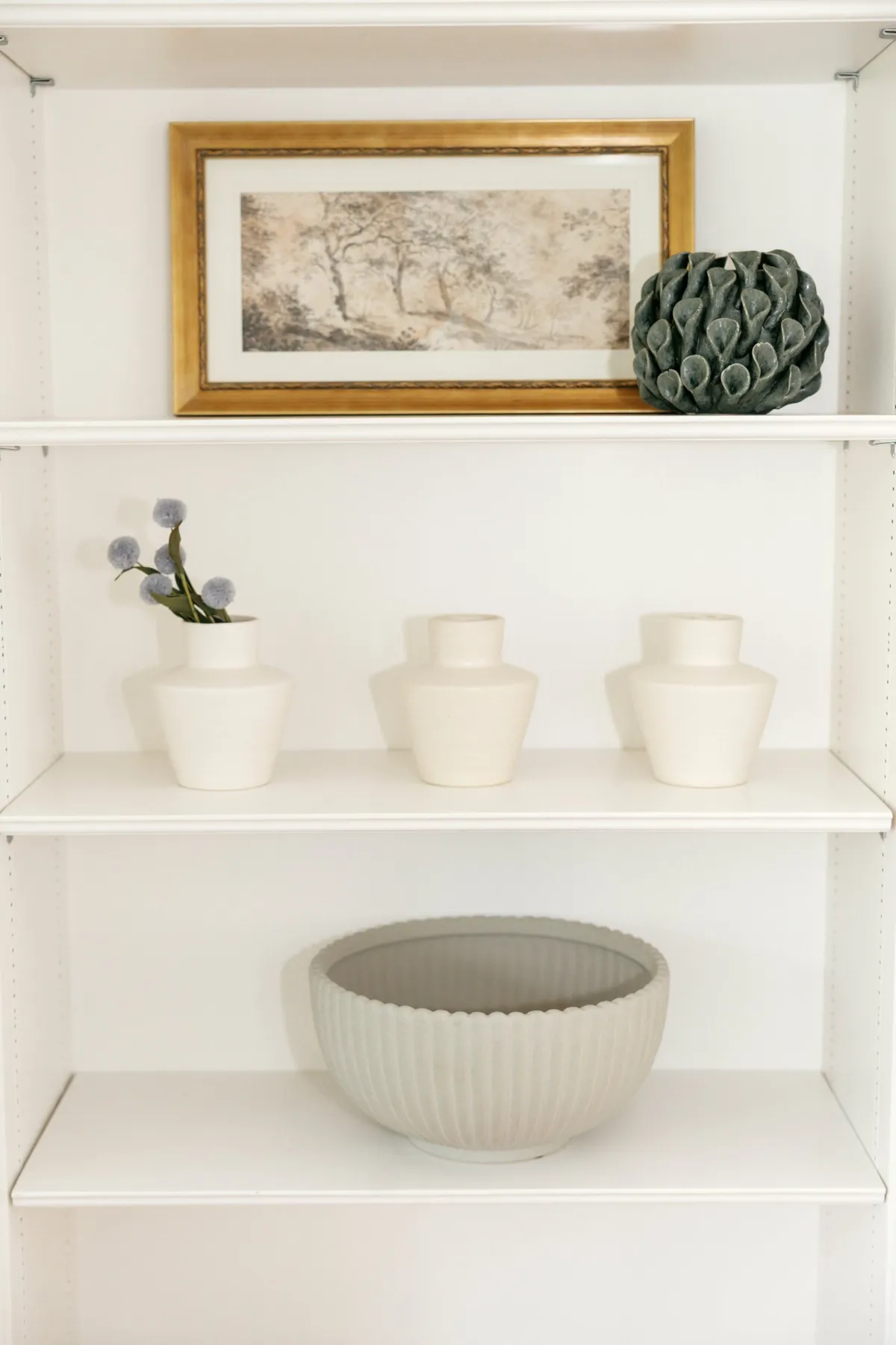 Framed landscape art, blue flowers, and a sculptural bowl on white shelves